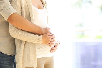 Ein Mann steht hinter einer Frau und umarmt sie. Die Hände des Mannes und der Frau liegen auf dem Bauch der Frau. Die Frau ist schwanger.