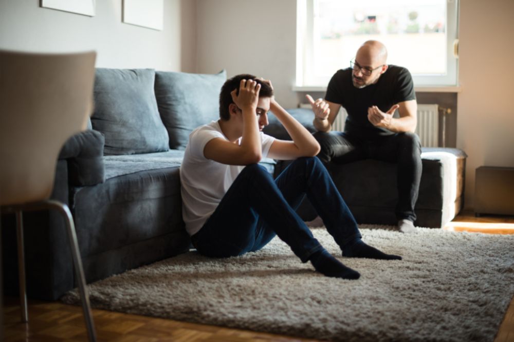 Junger Mann sitzt am Boden und hat seine Hände auf dem Kopf, er wirkt verzweifelt. Ein etwas älterer Mann sitzt auf einer Couch neben ihm und redet auf den jungen Mann ein.