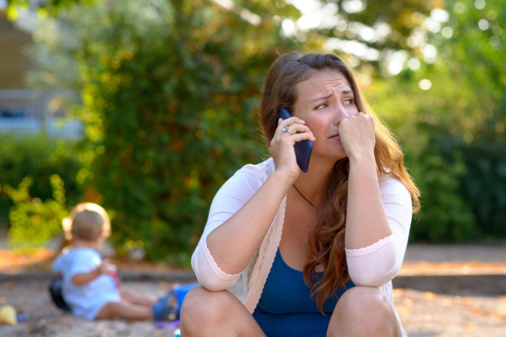 A woman is on her phone, she is crying. A baby is playing in the back.