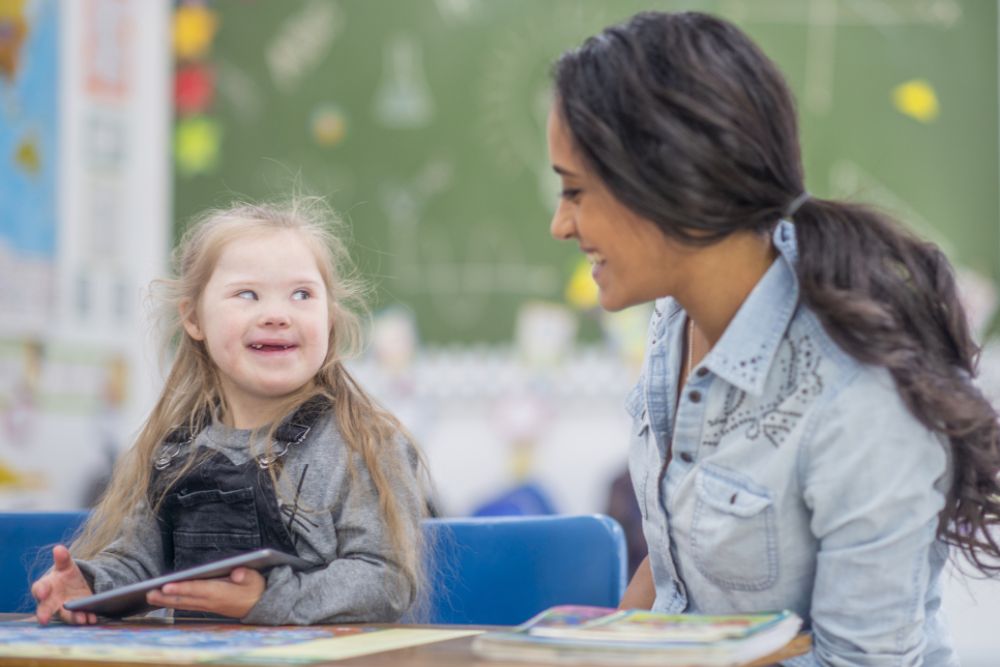 Eine Frau und ein Mädchen sitzen auf einem Tisch und lächeln sich an. Das Mädchen hat das Downsyndrom. 