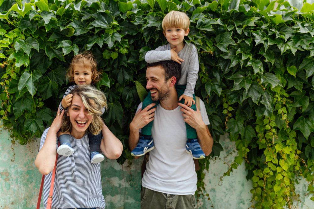 Eine Mutter und ein Vater tragen jeweils ein Kind auf ihren Schultern. Sie lachen herzlich.
