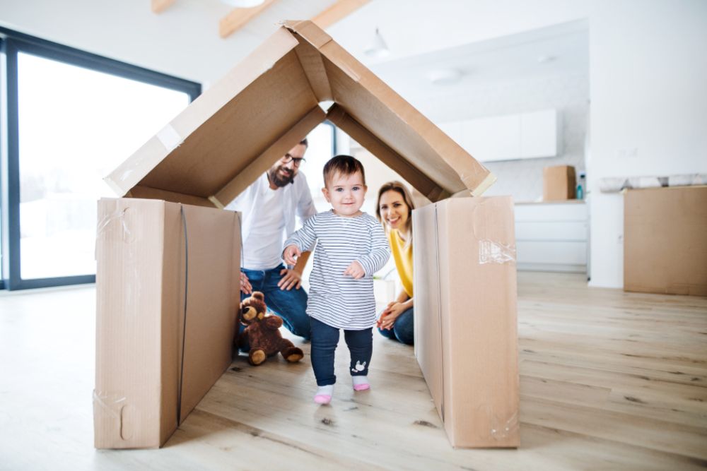 Familie mit Kind. Das Kind läuft durch ein Haus, welches aus Karton gebaut worden ist.