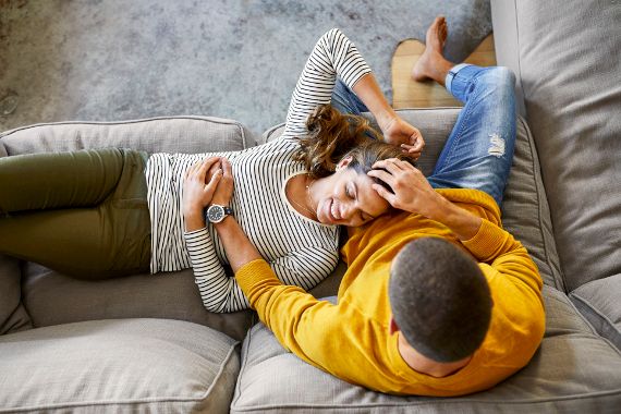 Ein Mann sitzt auf einem Sofa. Eine Frau liegt auf dem selben Sofa und liegt mit ihrem Kopf auf dem Schoß des Mannes. 