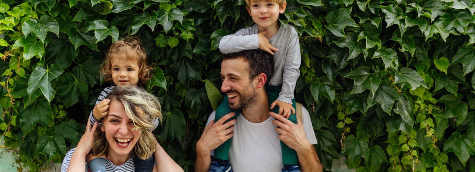  A woman and a man are standing in front of a wall. They each carry a child on their shoulders.